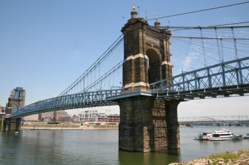 A historic suspension bridge spans a river, with city buildings visible in the background under a clear blue sky.