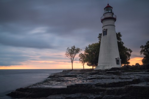 A lighthouse stands on rocky shore at dusk, with trees nearby and a colorful sunset in the background.