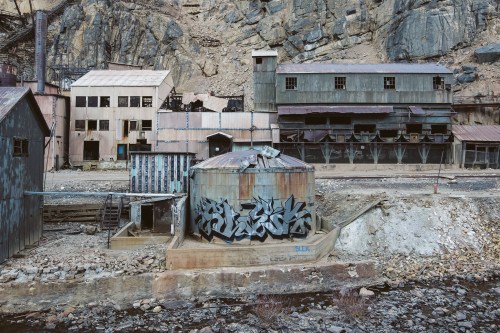 Abandoned industrial buildings and a graffiti-covered tank in a rocky, desolate landscape.