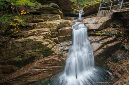 A serene waterfall cascades over rocky terrain, surrounded by lush greenery and autumn leaves.