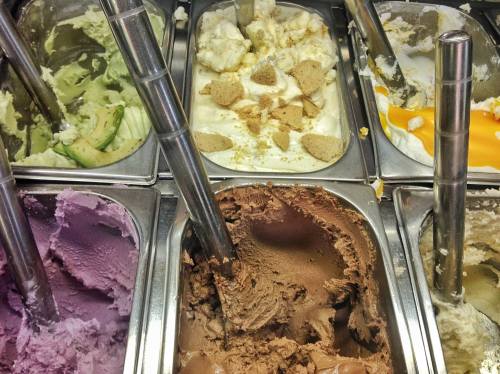 A variety of gelato flavors in metal containers, including green, white, purple, and brown, with scoops and toppings.
