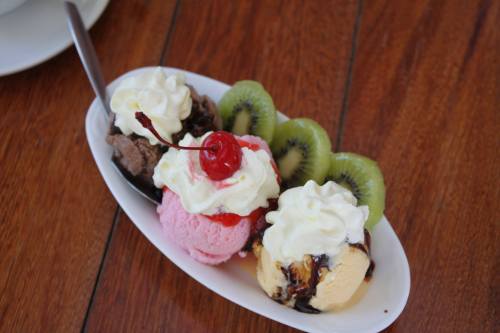 A dessert bowl with three scoops of ice cream (chocolate, strawberry, vanilla) topped with whipped cream, a cherry, and kiwi slices.
