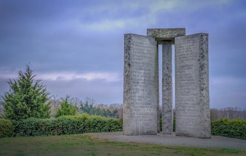 A large stone monument with engraved text, surrounded by greenery under a cloudy sky.