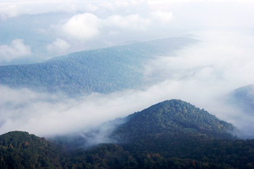A misty landscape of rolling green hills and valleys, partially obscured by clouds and fog.