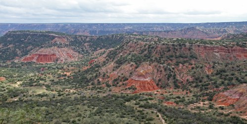 A panoramic view of colorful canyon landscapes with layered rock formations and green vegetation under a cloudy sky.