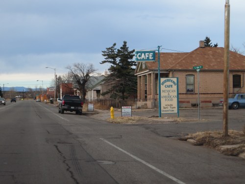 A roadside view of a Mexican café with a sign, surrounded by empty lots and a cloudy sky.