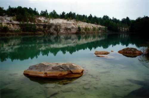 A serene lake with clear water reflecting rocky cliffs and trees, featuring a few large stones in the foreground.