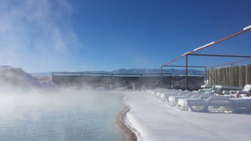 A steaming hot spring surrounded by snow, with lounge chairs and mountains in the background under a clear blue sky.