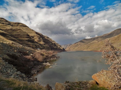 A serene river flows between rugged hills under a cloudy sky, with rocky banks and sparse vegetation.