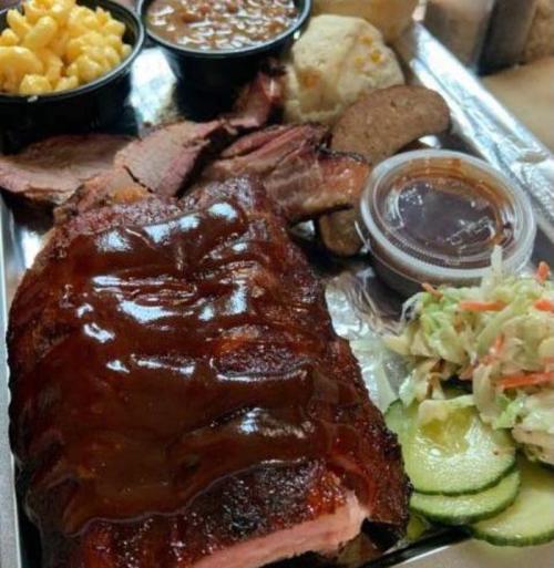 A barbecue platter featuring ribs, brisket, macaroni and cheese, baked beans, coleslaw, and pickles on a metal tray.