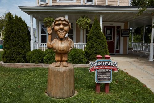 A wooden statue of a smiling man waving, in front of a house with green bushes and a sign about Wavin Walton.
