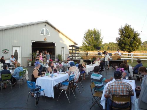 Outdoor gathering with tables set for dining, people socializing, and a speaker at a barn-like venue.