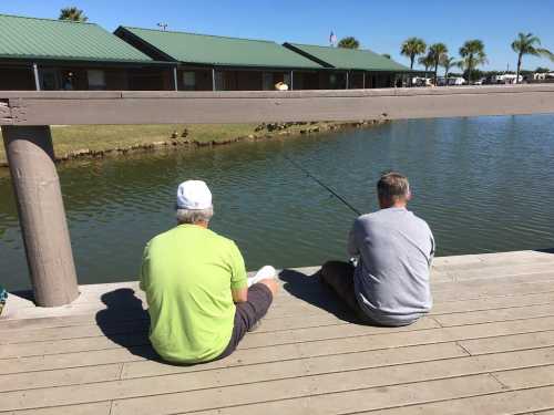 Two men sit on a wooden dock, fishing by a calm lake with palm trees and buildings in the background.