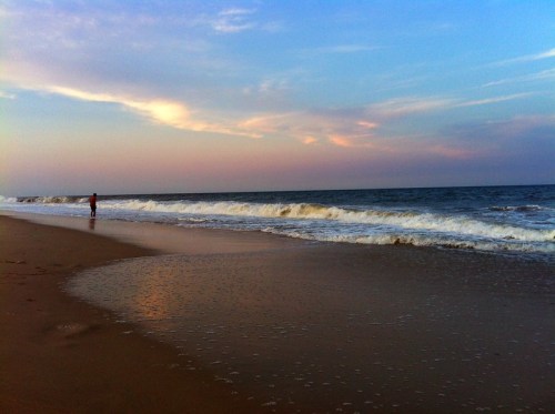 A person stands on a sandy beach, gazing at gentle waves under a colorful sky at sunset.