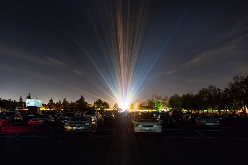A nighttime scene of a parking lot filled with cars, illuminated by colorful beams of light from a distant source.