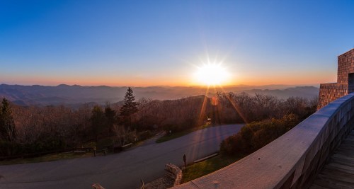 A panoramic view of a sunset over mountains, with vibrant colors and a clear sky, seen from a wooden overlook.