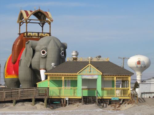A large elephant-shaped building next to a colorful beach house and a water tower against a clear blue sky.