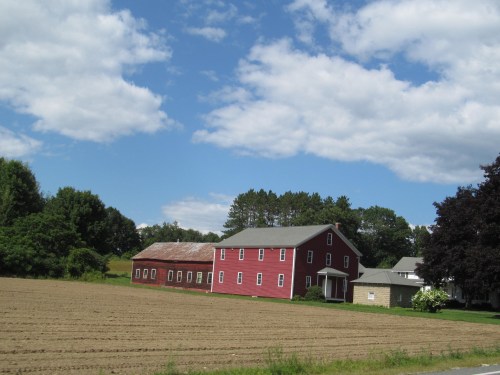 A red barn and farmhouse sit beside a plowed field under a blue sky with scattered clouds and green trees in the background.