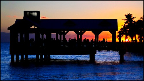 Silhouette of a pier with people against a vibrant sunset over the water, framed by palm trees.
