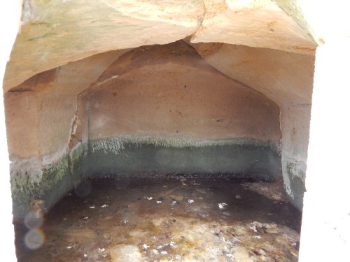 An empty, stone-walled cave with a damp floor and a curved ceiling, illuminated by natural light.