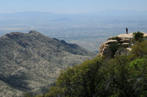 A person stands on a rocky outcrop overlooking a vast mountainous landscape and valley below.