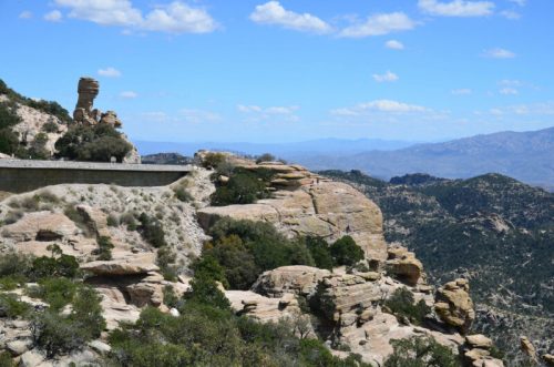 A scenic view of rocky cliffs and a winding road under a blue sky with scattered clouds. Mountains are visible in the distance.