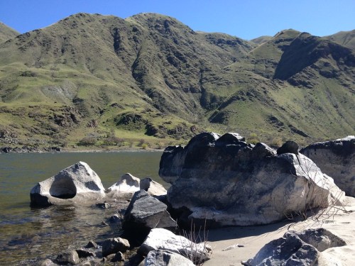 A serene landscape featuring rocky shores, calm water, and lush green mountains under a clear blue sky.