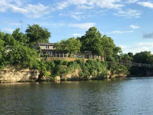 A serene river scene featuring a house surrounded by lush greenery and trees on a rocky bank under a blue sky.