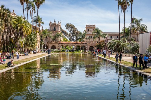 A scenic view of a palm-lined park with a reflecting pond and historic buildings in the background, bustling with visitors.