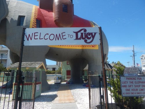 Sign reading "Welcome to Lucy" in front of a large, colorful elephant structure with a pathway and fences.