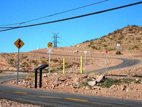 Winding road with traffic signs, power lines, and rocky terrain under a clear blue sky.