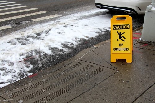 A yellow caution sign warns of icy conditions on a snowy sidewalk near a crosswalk.