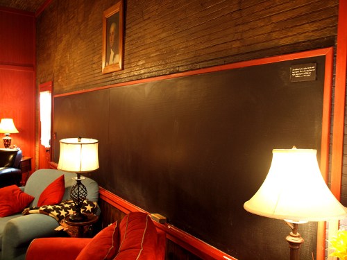 Cozy room with a large blackboard, two lamps, and colorful seating, featuring a portrait on the wall.