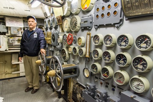 A man stands beside a control panel filled with gauges and levers in a submarine's control room.