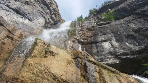 A cascading waterfall flows down a rocky cliff, surrounded by greenery and a clear blue sky.
