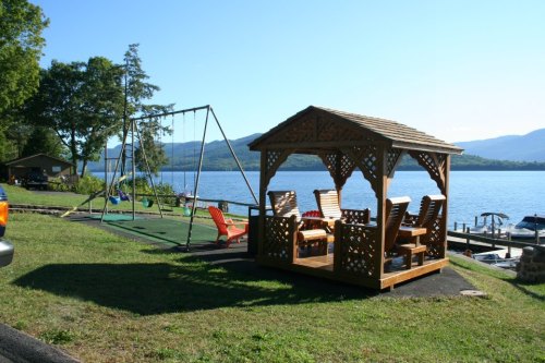 A wooden gazebo and swing set by a lake, with mountains in the background and a clear blue sky.