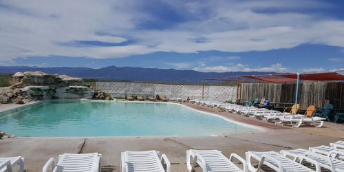 A serene outdoor pool surrounded by lounge chairs, with mountains and a blue sky in the background.