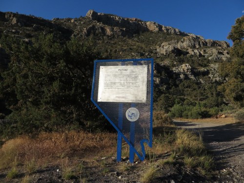 A blue informational sign about Potosi stands on a gravel path, surrounded by rocky hills and greenery.