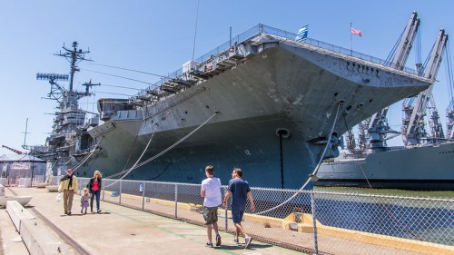 Visitors walk along the dock beside a large naval ship, with cranes and other vessels in the background.
