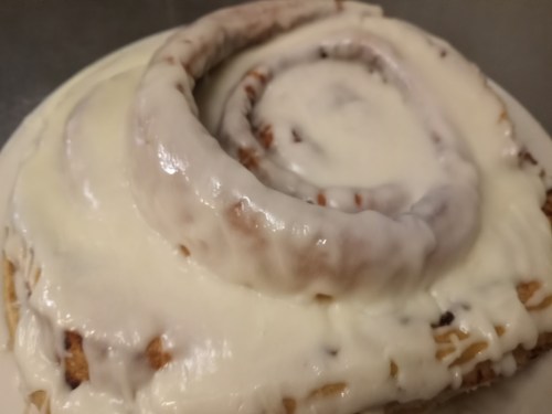 A close-up of a frosted cinnamon roll with a soft, spiraled shape and creamy icing on top.