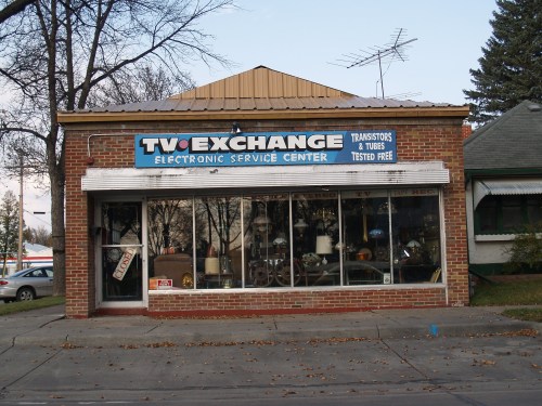 A brick storefront with a sign reading "Two Exchange Electronic Service Center" and large windows displaying various items.