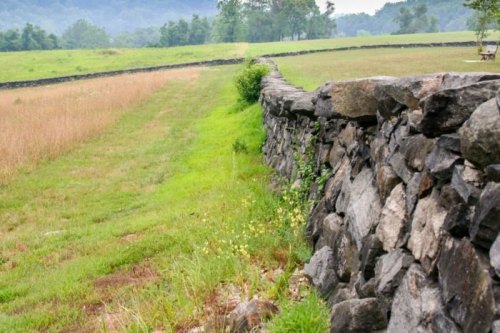 A stone wall runs along a grassy field, with trees and hills in the background under a cloudy sky.