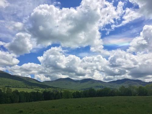 A scenic view of green mountains under a bright blue sky filled with fluffy white clouds and a hint of a rainbow.