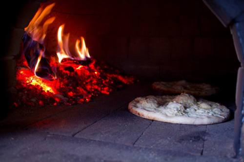 Two pizzas baking in a wood-fired oven, with flames and glowing embers in the background.