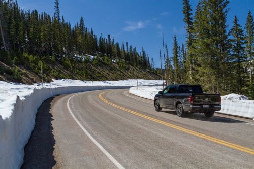A black truck drives along a winding road surrounded by snow and tall pine trees under a clear blue sky.