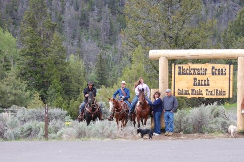 A group of people on horseback near a sign for Blackwater Creek Ranch, surrounded by trees and shrubs.