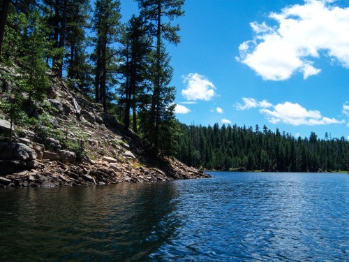 A serene lake surrounded by rocky shores and tall pine trees under a bright blue sky with fluffy clouds.