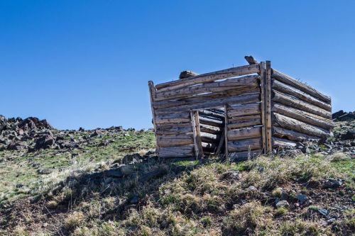 A weathered log cabin sits on a grassy hillside under a clear blue sky, surrounded by rocky terrain.