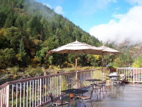 A scenic outdoor patio with tables and umbrellas, surrounded by lush green trees and a misty mountain backdrop.
