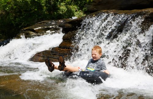 A young boy joyfully slides down a rocky waterfall on a black inner tube, splashing water around him.